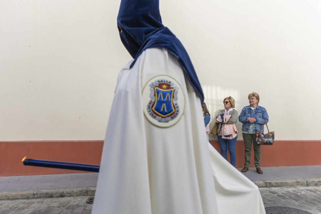 Un penitente de la Hermandad Dolor y Sacrificio pasa junto a dos señoras que esperan el paso de la procesiónón en El Puerto de Santa María en Cádiz, España durante la Semana Santa 2026 en la Bahía de Cádiz.