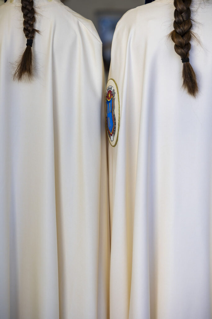 Foto vertical de dos chicas con sus trajes de penitentes vistas desde atrás ambas con el pelo recogido en una cola cayendo sobre la sotana blanca y rodeando el escudo de la Hermandad del Dolor y Sacrificio en El Puerto de Santa María durante la Semana Santa 2026 en la Bahía de Cádiz.