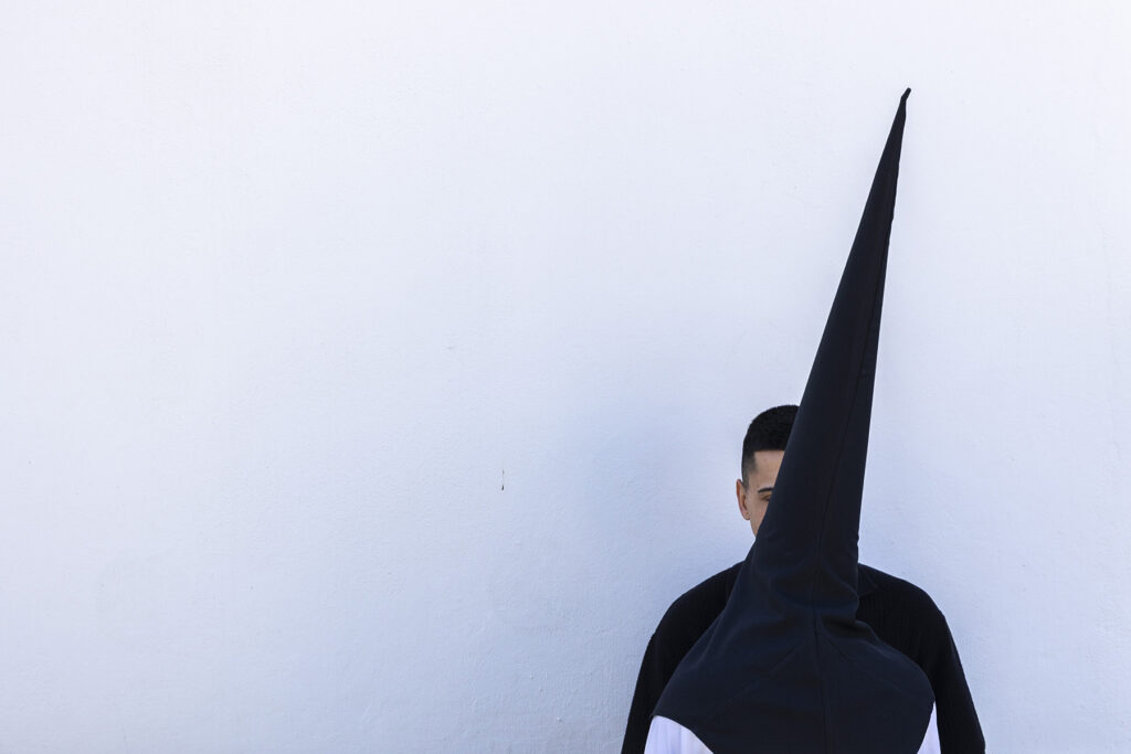 Foto de un penitente, con capirote negro y de espaldas, hablando con otra persona ambos contra una pared blanca, de Hermandad del Perdón en San Fernando, Cádiz, España durante la Semana Santa de 2026.