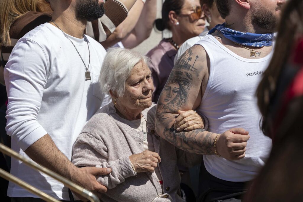 Un mujer rodeada y protegida por sus nietos, cargadores de la Hermandad de El Perdón, observa la procesión a su paso por una barriada de San Fernando, Cádiz, España durante la Semana Santa 2026 en la Bahía de Cádiz.