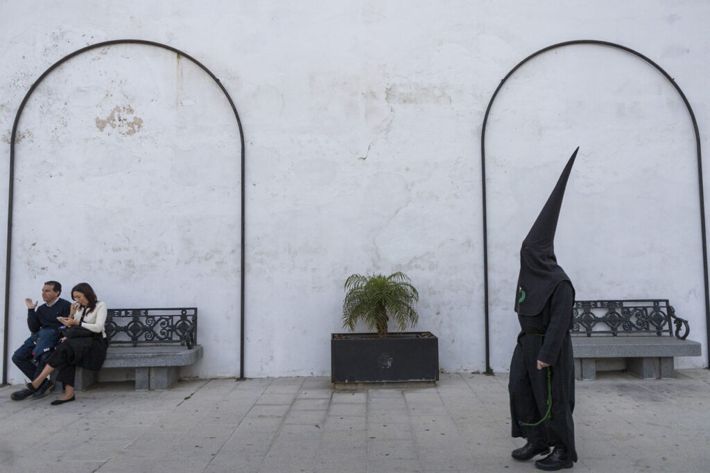 Un penitente vestido totalmente de negro pasa junto una pareja sentada en una plaza en San Fernando, Cádiz, España, momentos antes de la salida de los pasos de la Hermandad de La Expiración durante la Semana Santa 2026 en la Bahía de Cádiz.