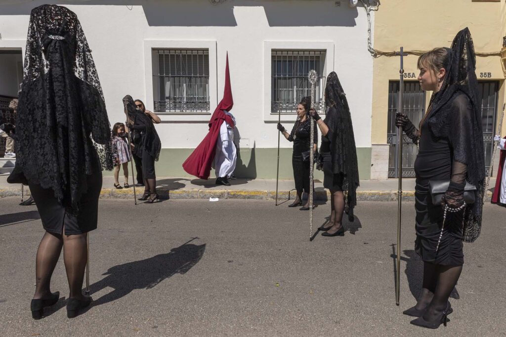 Un penitente de la Hermandad de Las Tres Caídas pasa junto a un grupo de mujeres ataviadas con mantilla y peineta durante la Semana Santa 2026 en la Bahía de Cádiz en San Fernando, España.